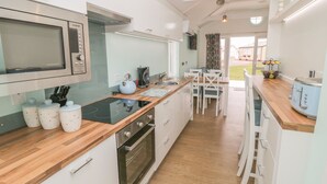 A modern kitchen with a microwave, kettle, and stove. There is a dining area with chairs and a table. The kitchen has white cabinets and a wooden countertop. - Shore Thing 280 (Bridlington)