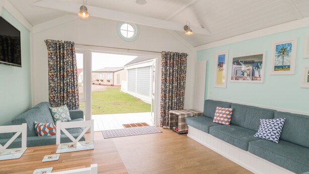 A cozy living room with a wooden dining table, a blue sofa, and a white chair. There is a small table with a lamp and a window with curtains. - Shore Thing 280 (Bridlington)