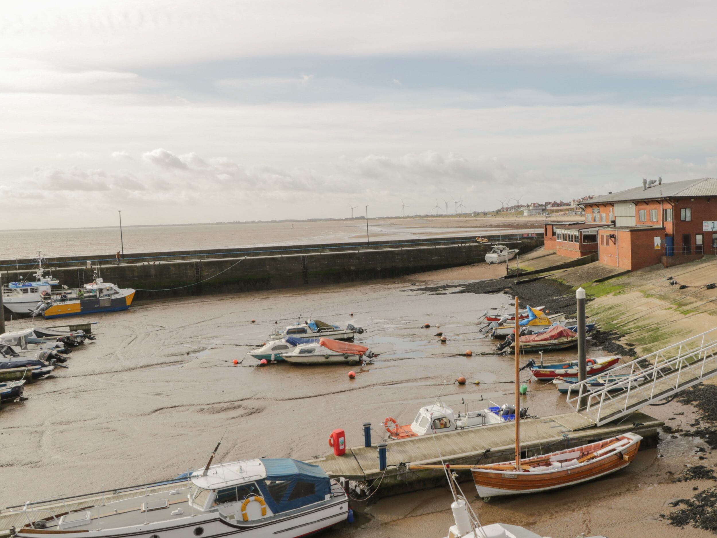 A coastal marina with boats docked along the shore, a sandy beach, and a building with a red roof in the background.