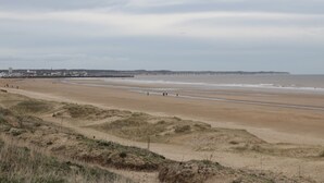 A wide sandy beach with gentle waves, a distant shoreline with buildings, and overcast sky. - Shore Thing 280 (Bridlington)
