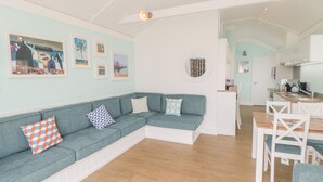 A modern living room with a teal sofa, white walls, and a wooden floor. There is a dining area with white chairs and a kitchen in the background. - Shore Thing 280 (Bridlington)