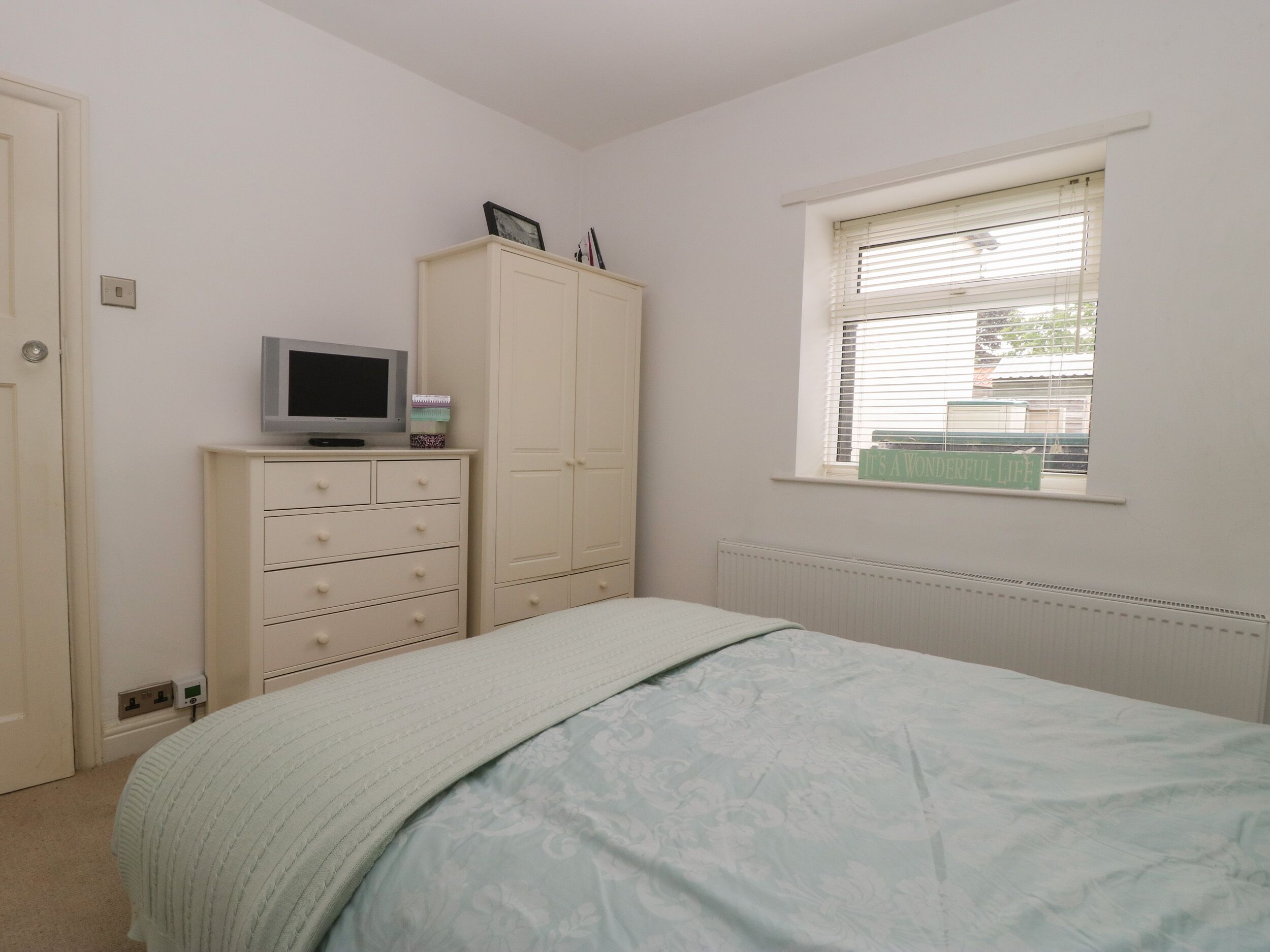 A bedroom with a bed, a dresser, a television on top, and a window with blinds.
