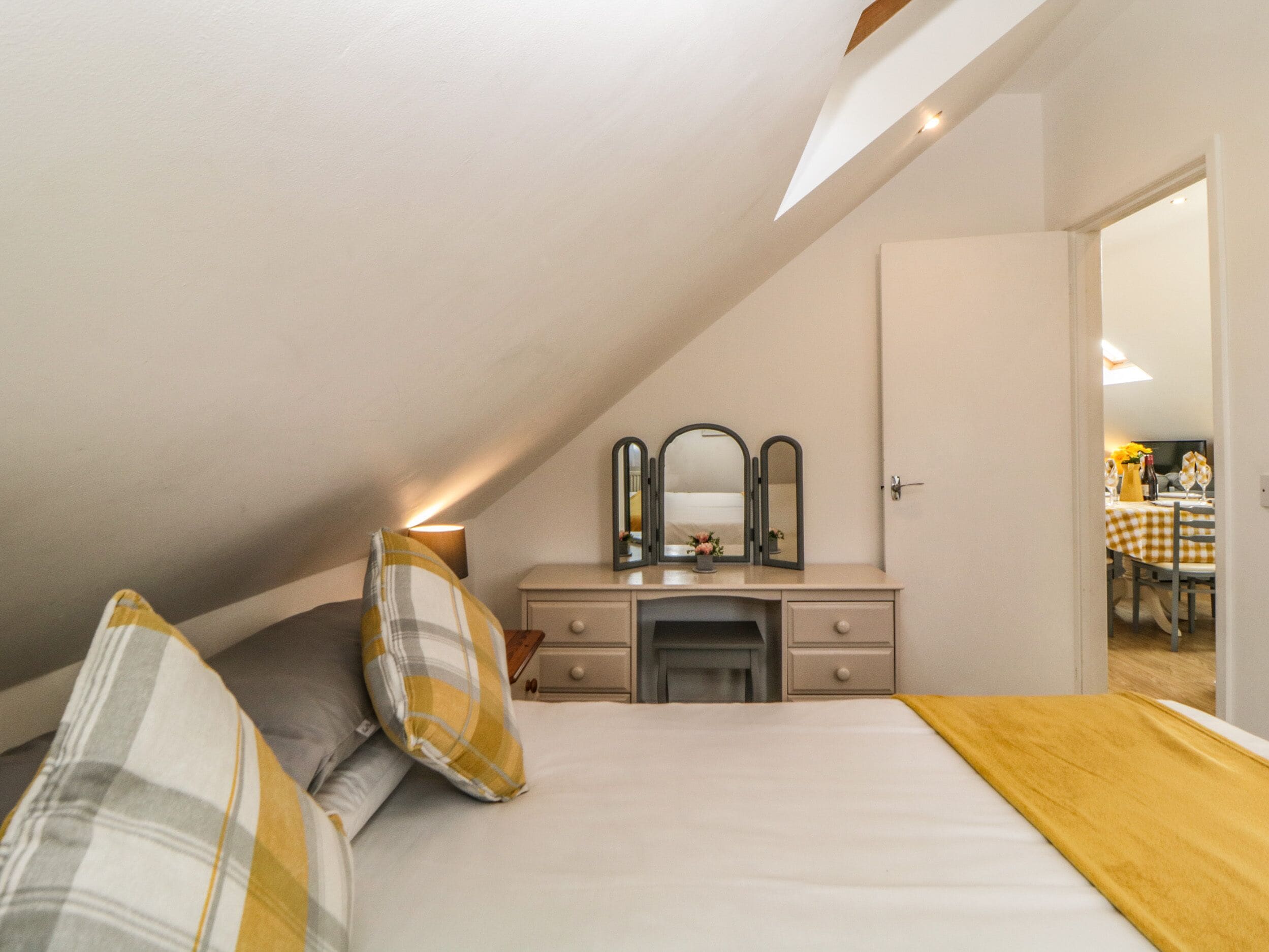 A bedroom with a sloped ceiling, a bed with a white and yellow checkered bedspread, a bedside table with a mirror, and a dresser.