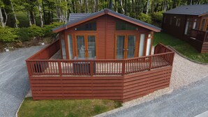 A wooden cabin with a balcony, surrounded by gravel and grass, with trees in the background. - Lodge 11 (Tavistock)