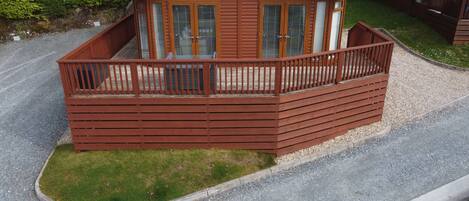 A wooden cabin with a balcony, surrounded by gravel and grass, with trees in the background.