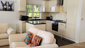 A modern kitchen with white cabinets, a black countertop, and a stainless steel range hood. There is a dining area with a beige sofa and colorful cushions. - Lodge 11 (Tavistock)