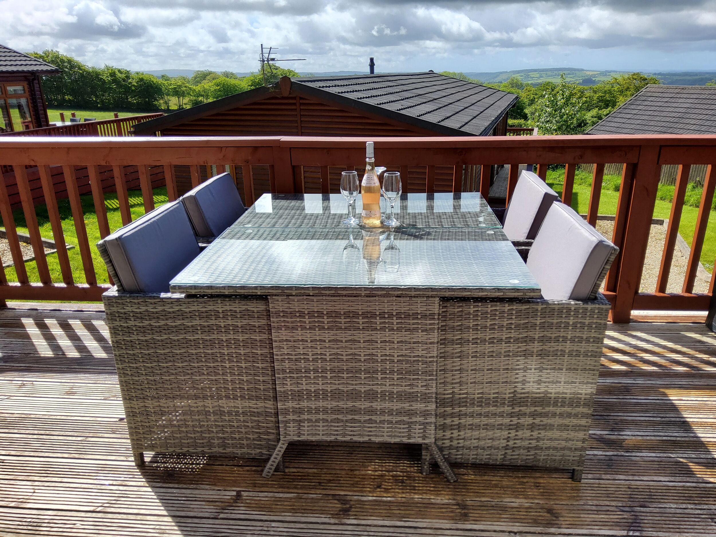Een rieten tafel met een glazen blad, twee wijnglazen en een fles op een terras met een houten balustrade en uitzicht op een groen landschap.