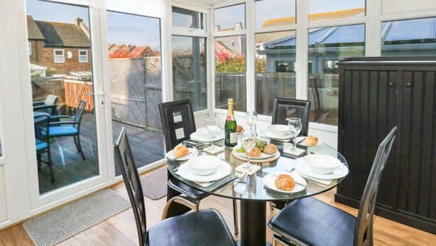 Een zonnige eetruimte met een glazen tafel, stoelen en uitzicht op een tuin en omliggende huizen.