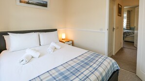 Een net opgemaakt bed met witte lakens en een blauw geruit plaid, een bedlamp en een ingelijste foto aan de muur.