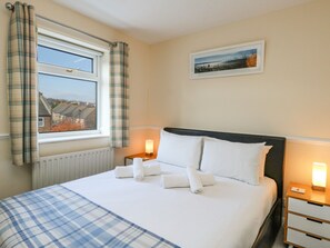 A neatly made bed with white linens and rolled towels, a window with plaid curtains, a bedside table with a lamp, and a framed picture on the wall.