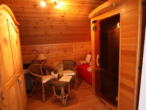 A wooden cabin interior with a dining table, two wicker chairs, a red sofa, and a door leading to a sauna.