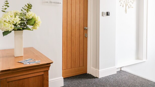 A reception area with a wooden door labeled 'Dovedale', a white vase with flowers, and a small table with brochures. - Dovedale (Ashbourne)