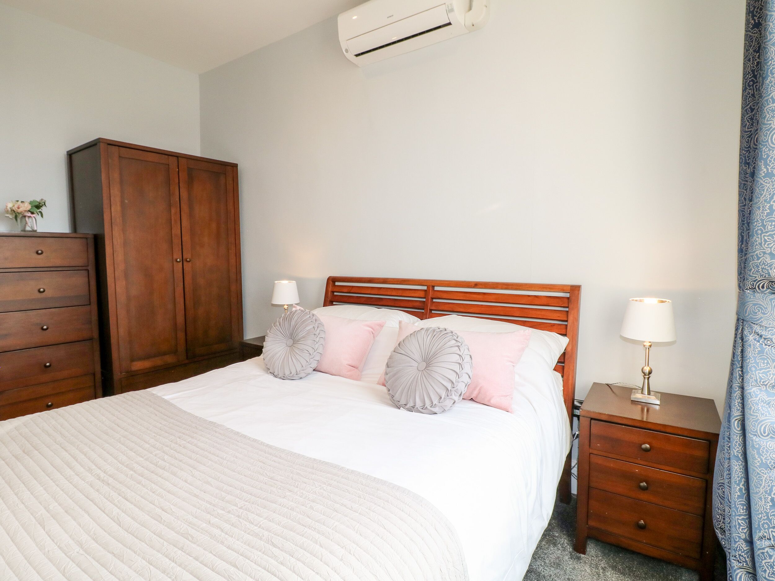 Een netjes opgemaakt bed met een witte deken, twee nachtkastjes met lampen, een houten kledingkast en een airconditioning op de muur.