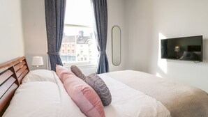 A hotel room with a bed, a TV, a window with curtains, and a view of buildings outside. - Dovedale (Ashbourne)