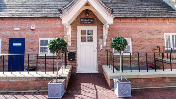 A brick building with a white door and a sign reading "Derbyshire House". There are two potted plants on either side of the entrance. - Milldale (Ashbourne)