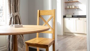 A wooden dining table with a chair, a window with curtains, and a kitchen area with white cabinets and shelves. - Milldale (Ashbourne)
