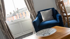 A room with a blue armchair, a wooden side table, and a window with patterned curtains. - Milldale (Ashbourne)