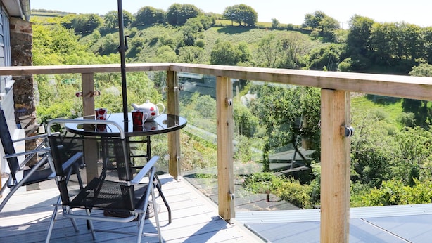 A balcony with a table, chairs, and an umbrella, overlooking a lush green landscape. - Higher Bumsley Barn (Barnstaple)