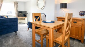 A modern living room with a wooden dining table, two chairs, a blue sofa, a TV, and a lamp. - Manifold Dale (Ashbourne)