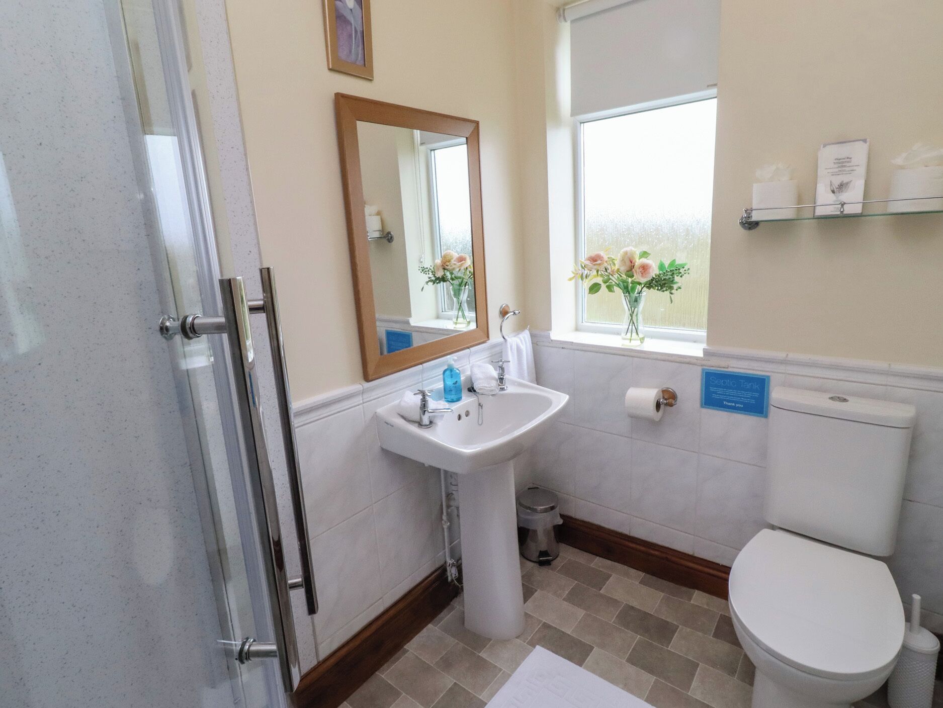 A bathroom with a glass shower enclosure, a sink with a mirror, a toilet, and a window with a view of flowers.
