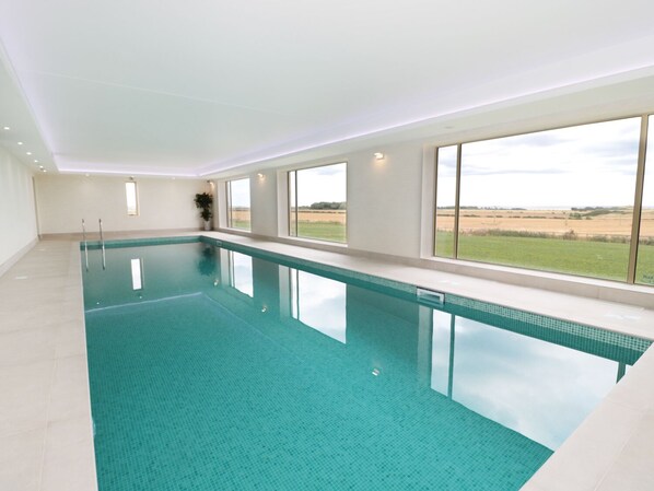 A large indoor swimming pool with clear blue water, surrounded by large windows offering views of a grassy landscape.