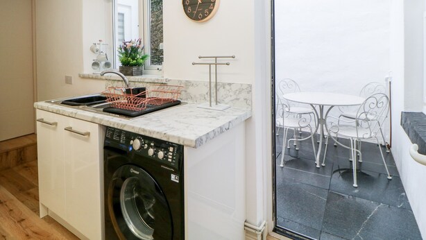 A compact kitchen with a washing machine, a sink with a dish rack, and a dining area with a table and chairs. - Cove Cottage (Torquay)