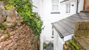 A narrow alleyway with a stone wall on one side and a white building with a black roof on the other. There is a small table and chairs outside the building. - Cove Cottage (Torquay)