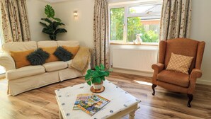 Una sala de estar con un sofá color crema, un sillón marrón, una mesa de centro con revistas, una planta grande y una ventana con cortinas.
