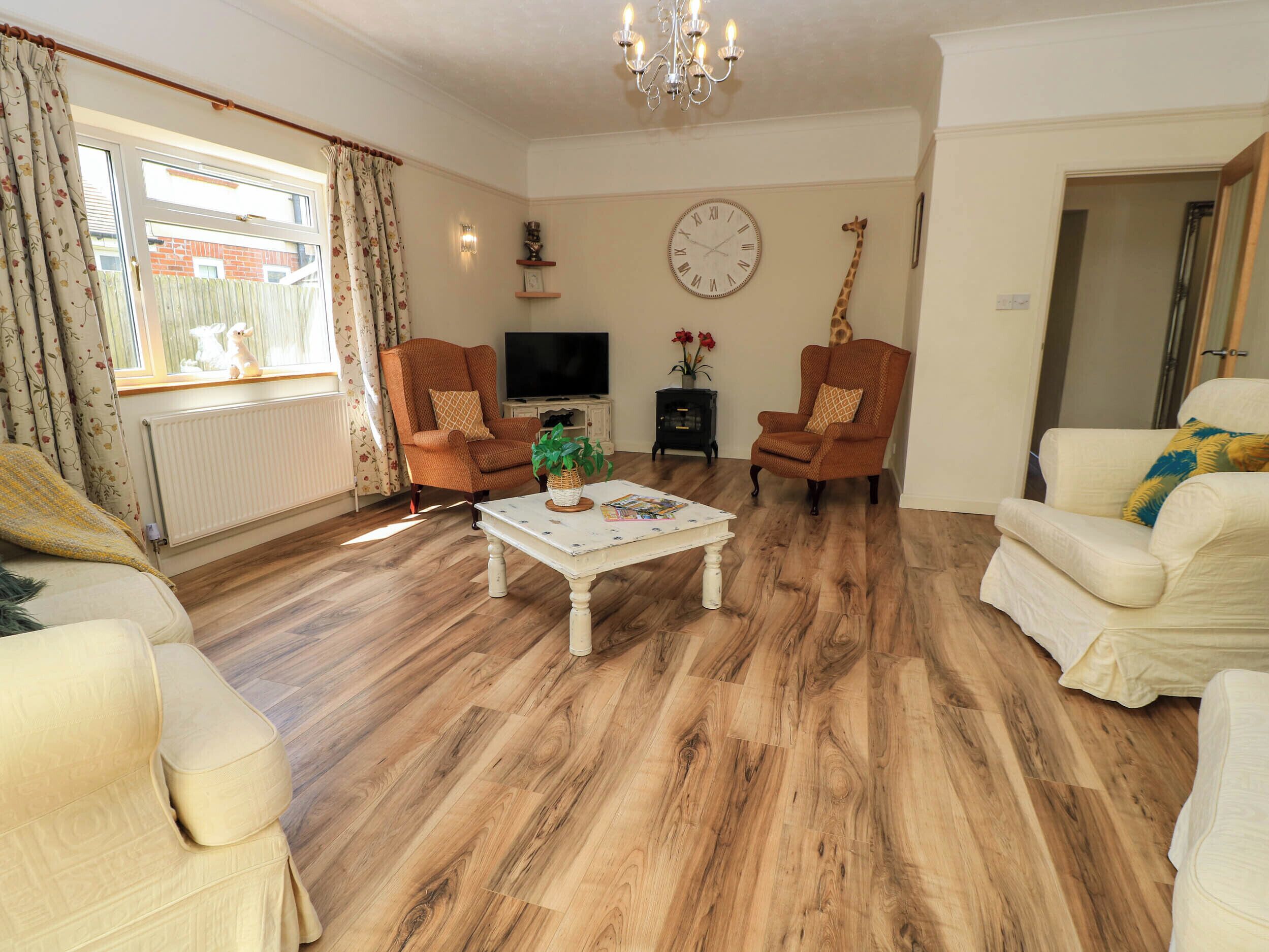 A living room with a wooden floor, two armchairs, a sofa, a coffee table, a TV, and a wall clock.