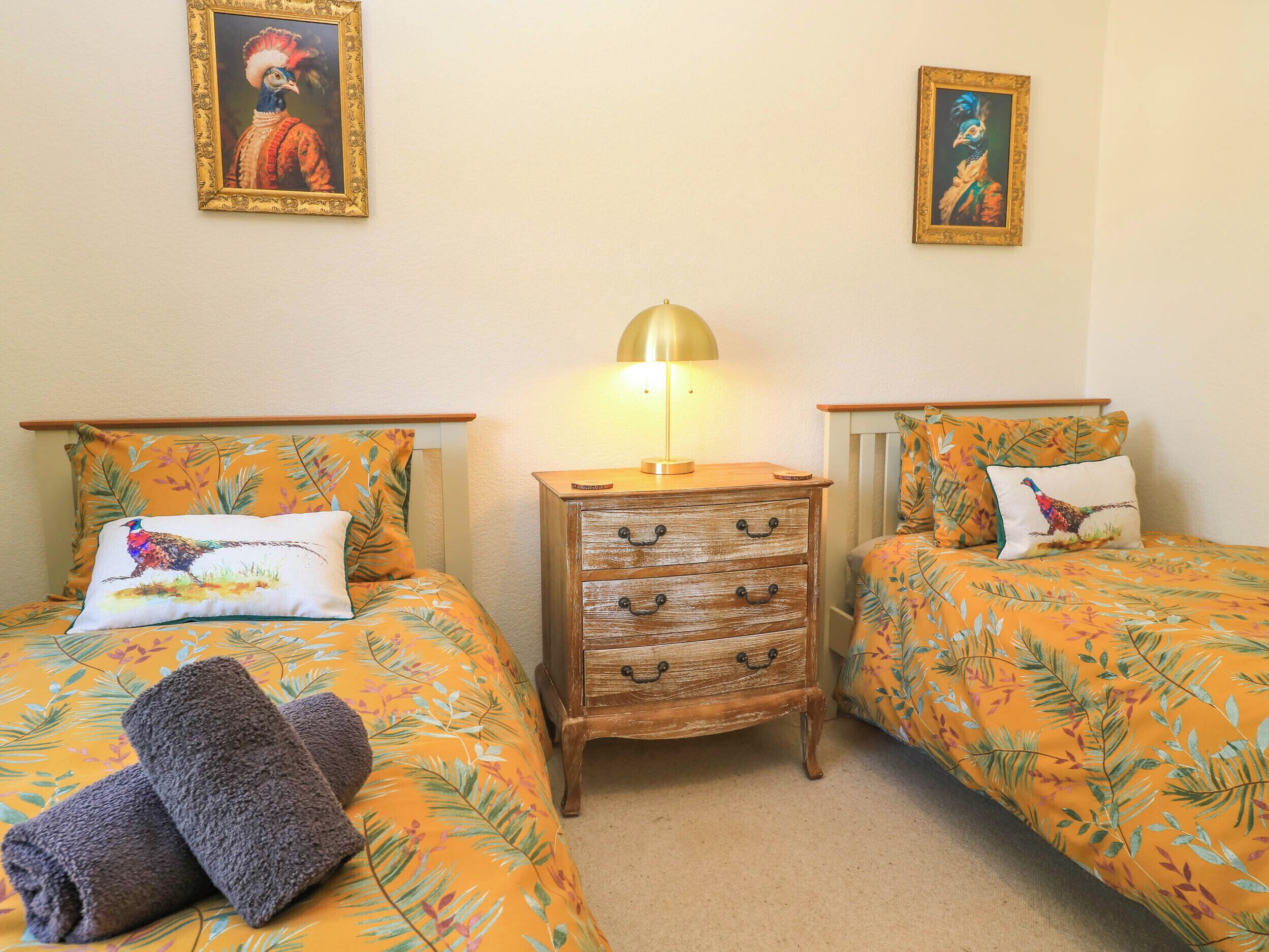 Una habitación con dos camas, cada una con colcha y almohadas a juego con motivos florales. Entre las camas hay una mesita de noche de madera con una lámpara.