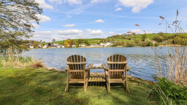 Two wooden Adirondack chairs facing a lake with a grassy foreground and houses in the background. - Clerk Beck Cottage (Ulverston)