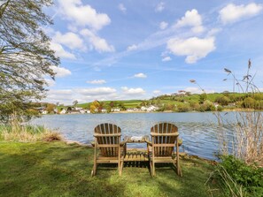 Duas cadeiras de madeira tipo Adirondack viradas para um lago, com um gramado à frente e casas ao fundo.