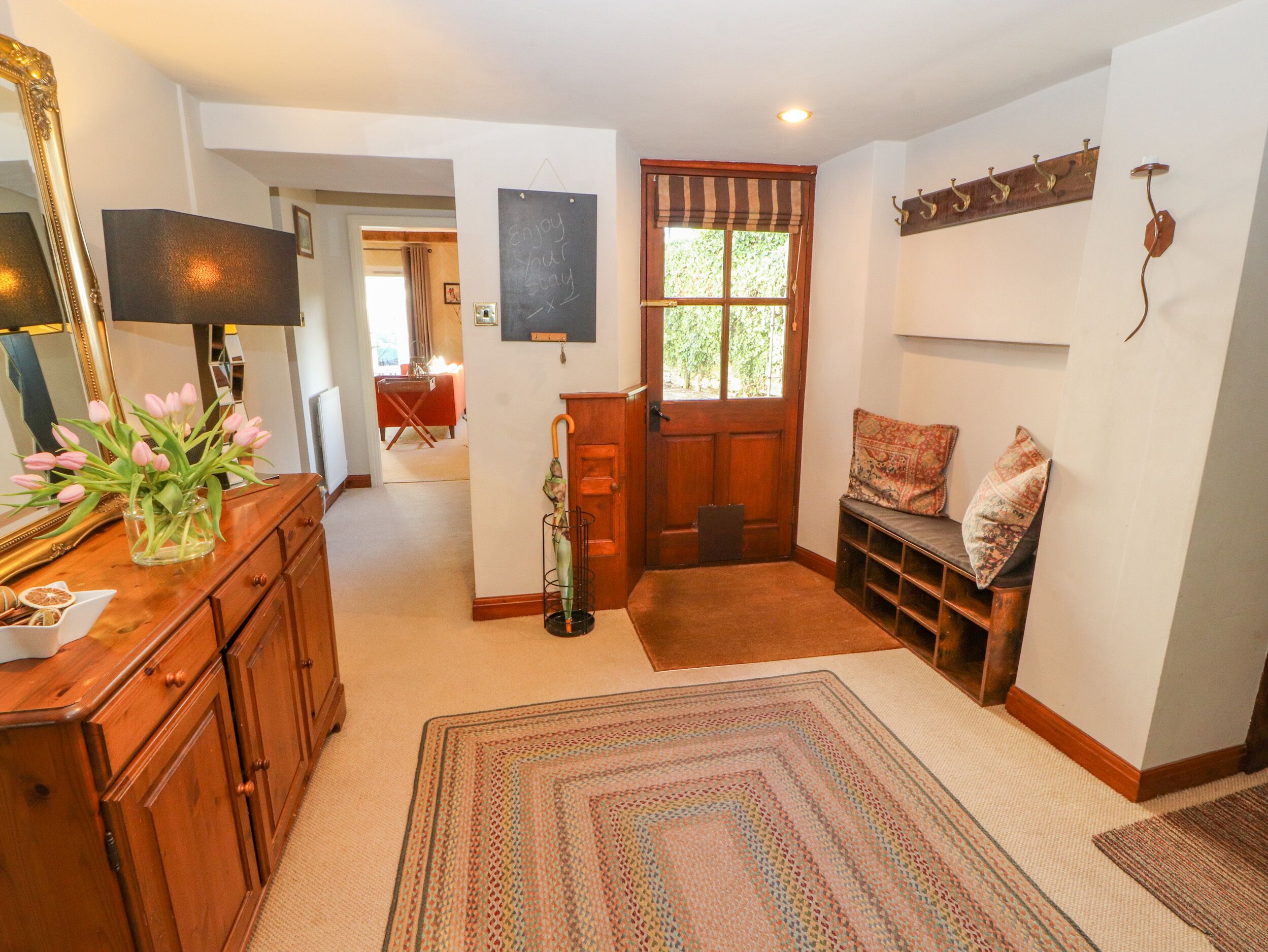 A cozy room with a wooden dresser, a mirror, a vase of pink tulips, a bench with cushions, and a door with a window.
