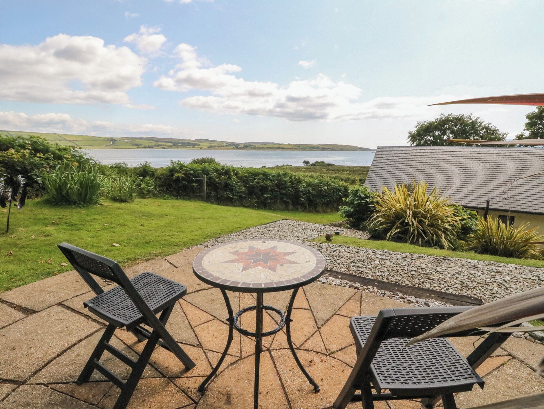 Eine Terrasse mit Tisch und Stühlen, Blick auf einen See und eine Rasenfläche mit einer Hecke.