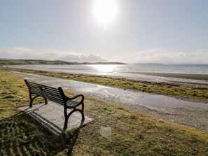 A bench by a beach with a view of the sea and distant hills.
