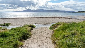 Een zandpad langs het strand dat uitkomt bij een rustige zee, met bergen in de verte.