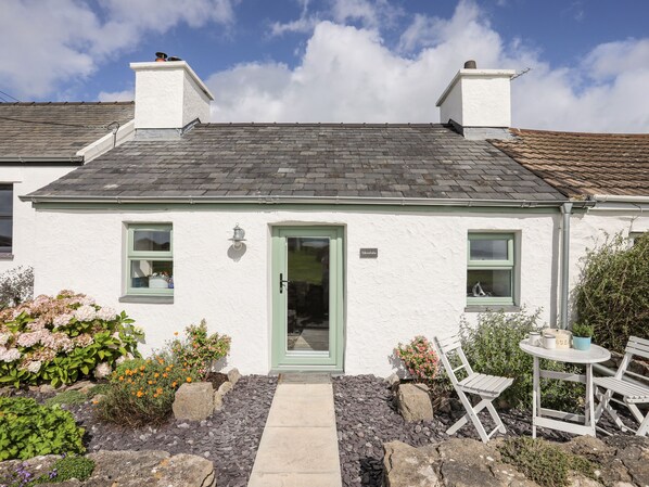 A white cottage with a green door, two windows, and a small garden with a table and chairs. - Minnehaha (Moelfre)