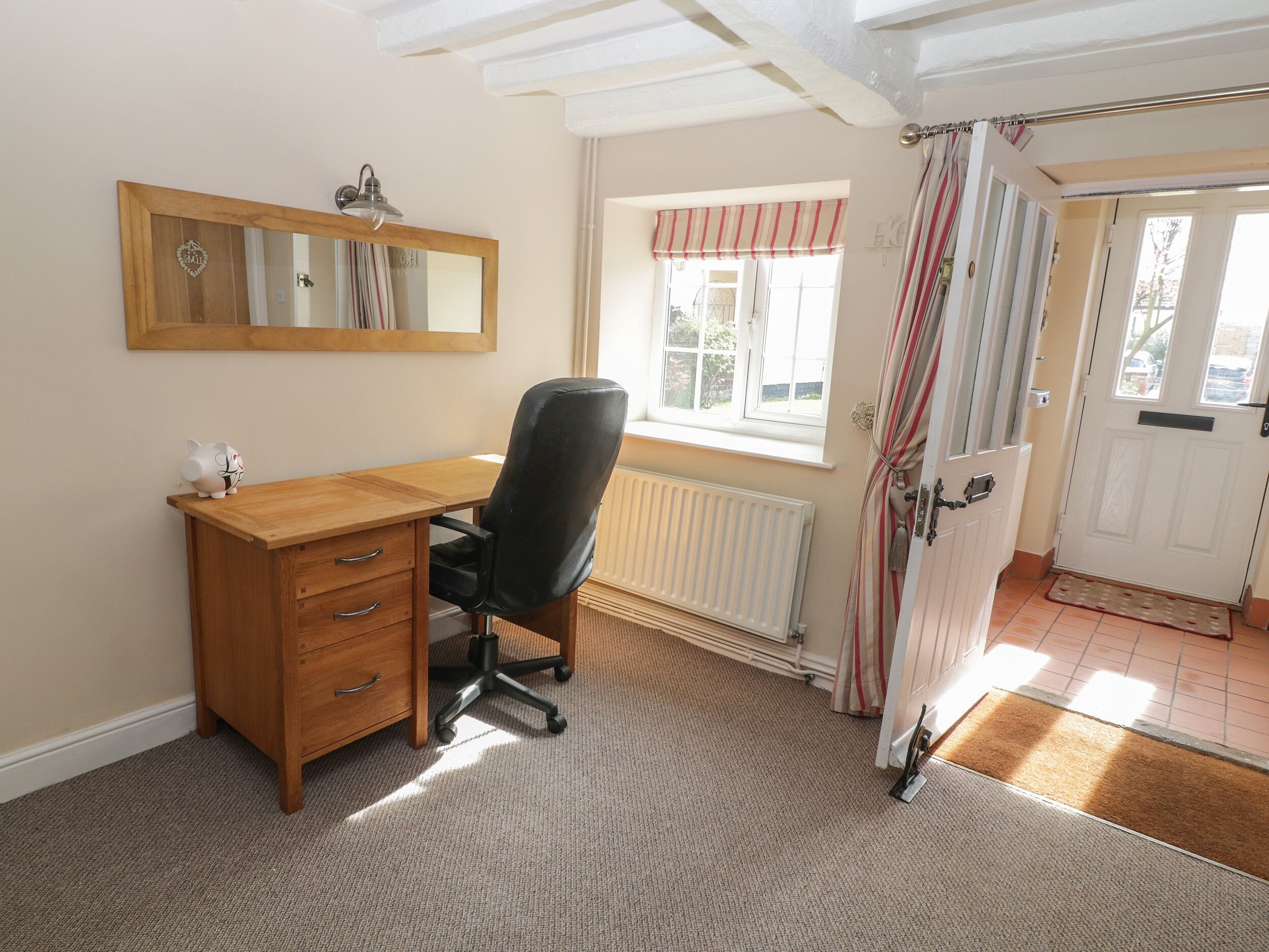 A room with a wooden desk, a black office chair, a radiator, a window with curtains, and a door with a striped curtain.