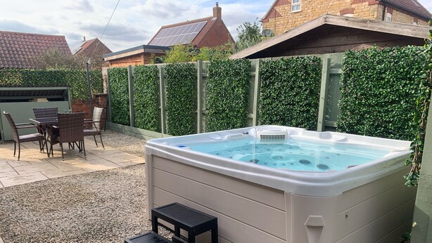 A hot tub in a backyard with a patio table and chairs, surrounded by a hedge and a gravel area. - Farriers Cottage (Lincoln)