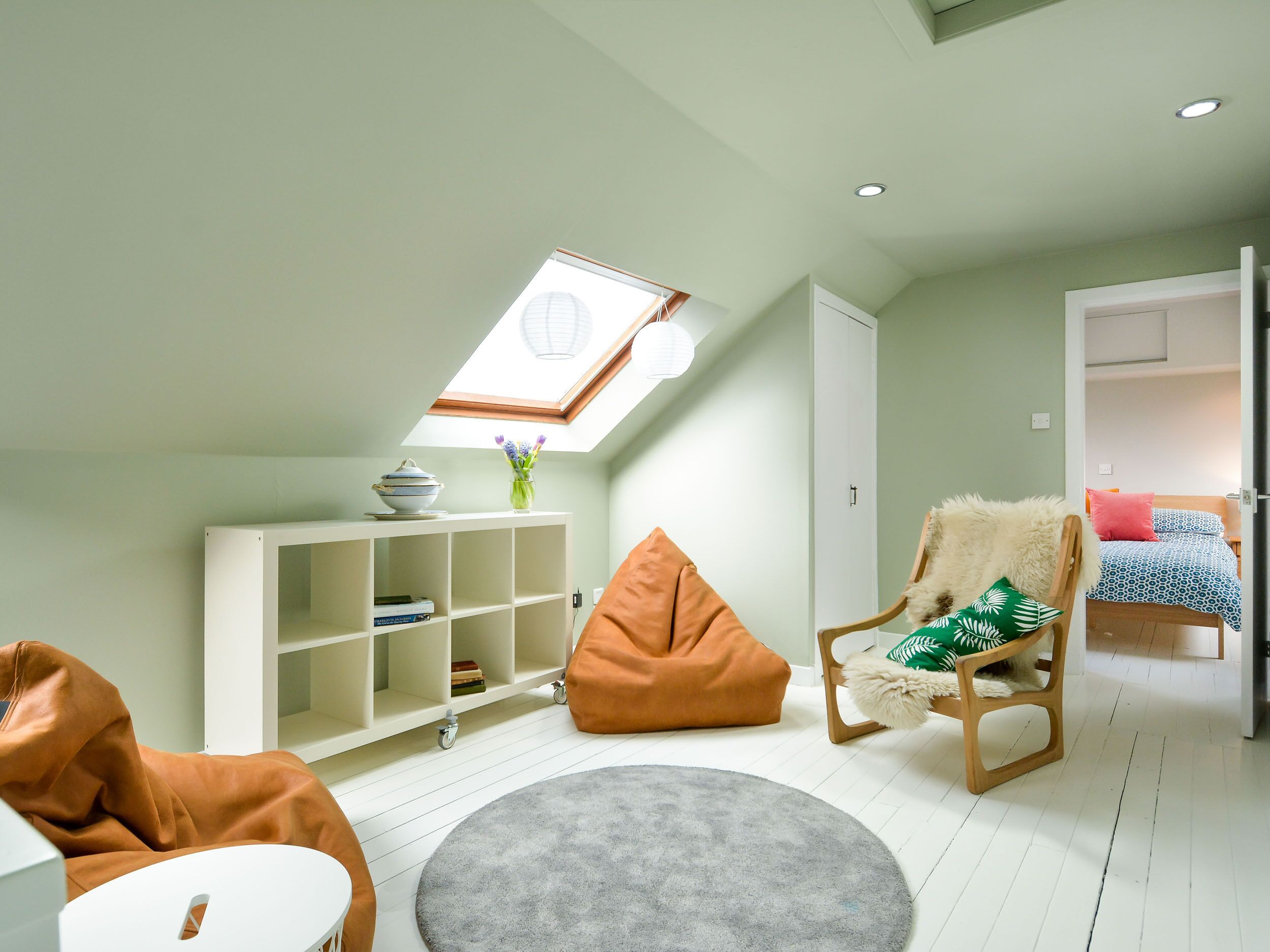 A cozy loft bedroom with a skylight, a white wardrobe, a wooden chair with a fluffy cushion, and a bed with patterned bedding.