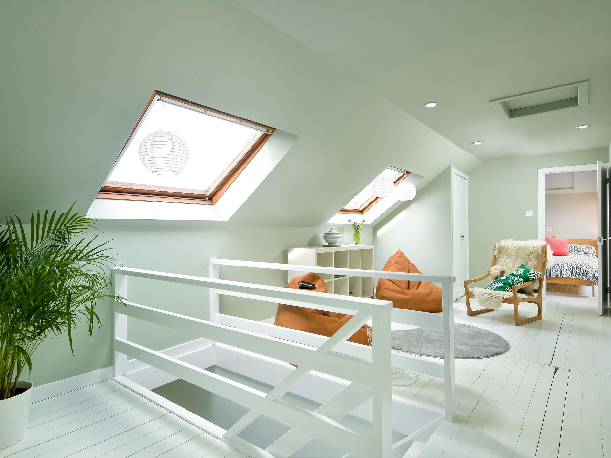 A modern loft bedroom with skylights, a white bed, a chair with a fluffy cushion, and a potted plant.