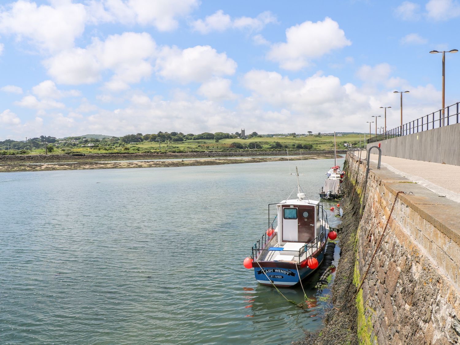 Un barco llamado 'Lady Wendy' atracado en un muelle con un muro de piedra y un paisaje con césped al fondo.