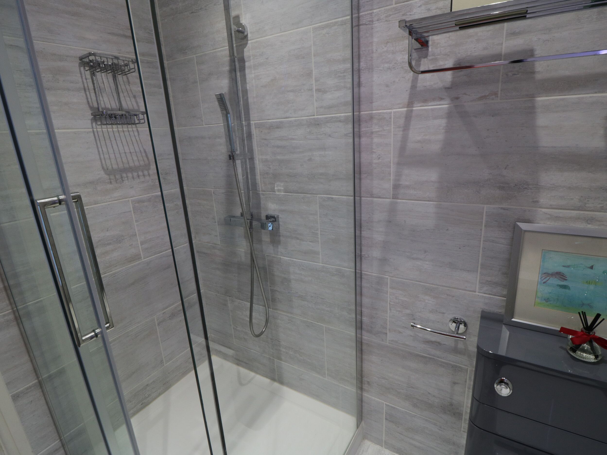 Un baño moderno con cabina de ducha de vidrio, azulejos grises, un cuadro en el tocador y un perchero.