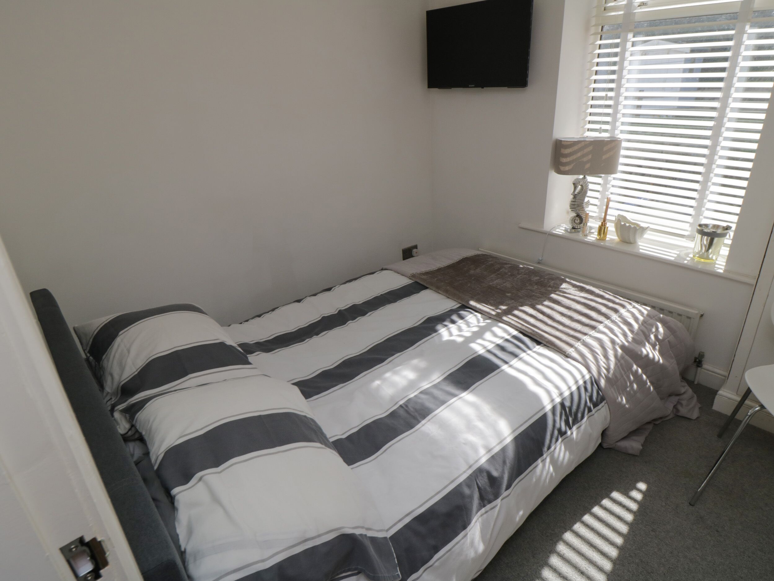A bedroom with a bed, a TV mounted on the wall, a window with blinds, and a bedside table with a lamp.