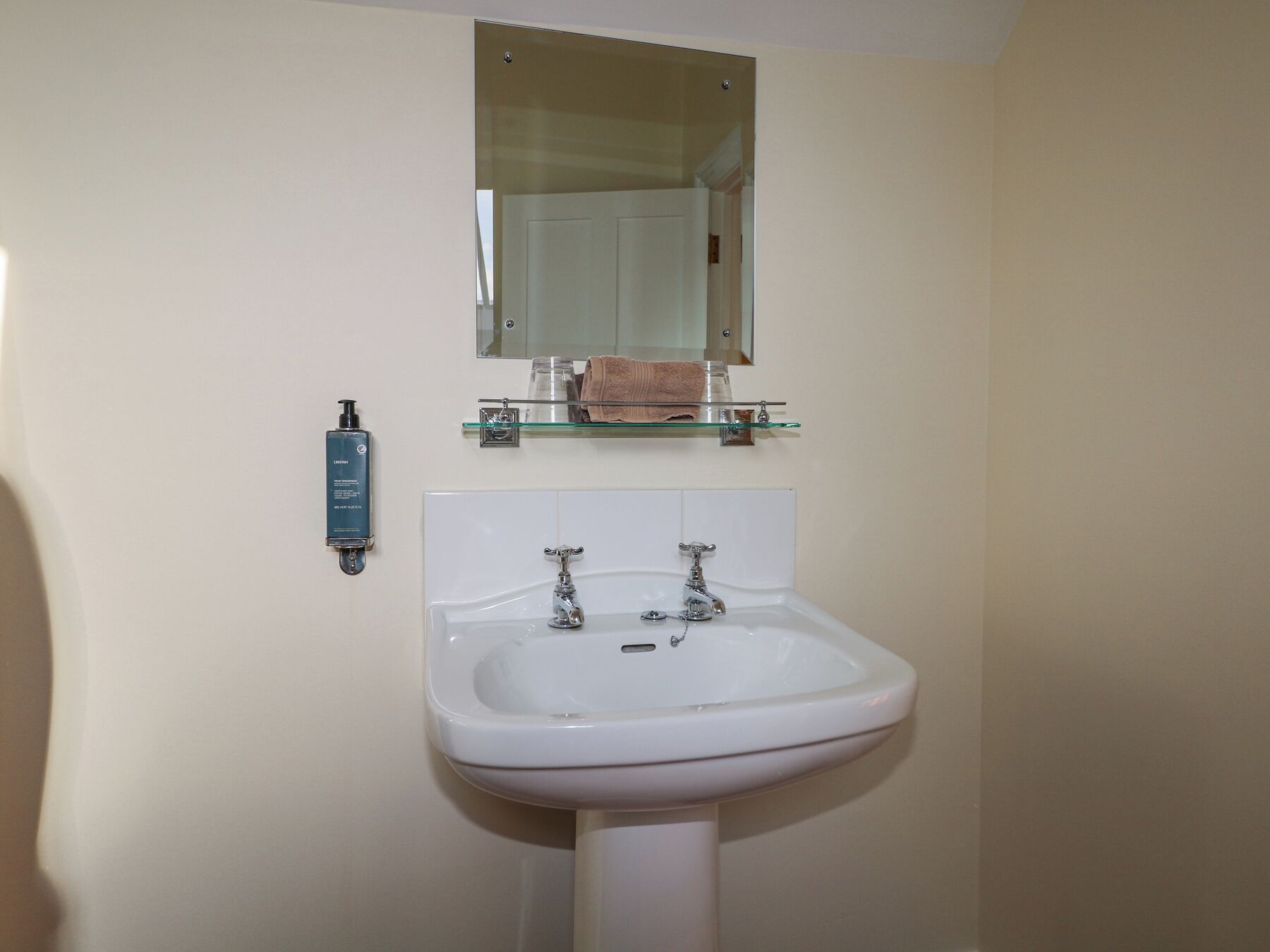 A bathroom with a white sink, two faucets, a glass shelf with a towel, and a mirror above the sink.