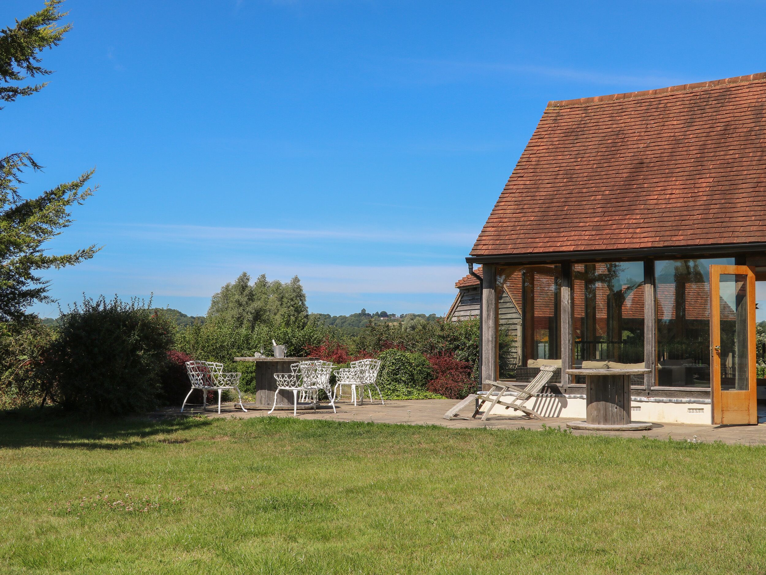 Une maison avec un toit recouvert de tuiles rouges, une véranda fermée par des vitres et une cour verdoyante aménagée avec du mobilier d’extérieur.