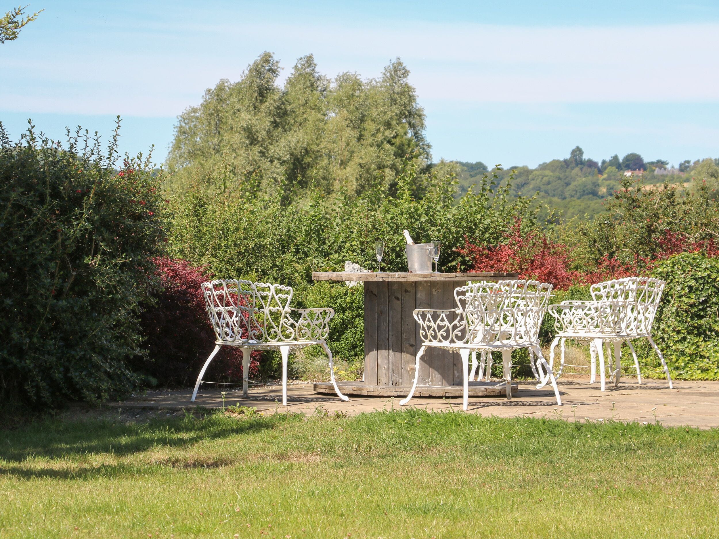A patio with white metal chairs and a table, surrounded by lush greenery and trees.
