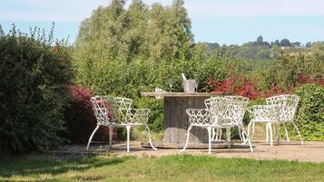 A patio with white metal chairs and a table, surrounded by lush greenery and trees.