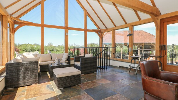 A covered patio area with wicker furniture, a stone floor, and large windows offering a view of the surrounding landscape. - Groomes Country House (Bordon)