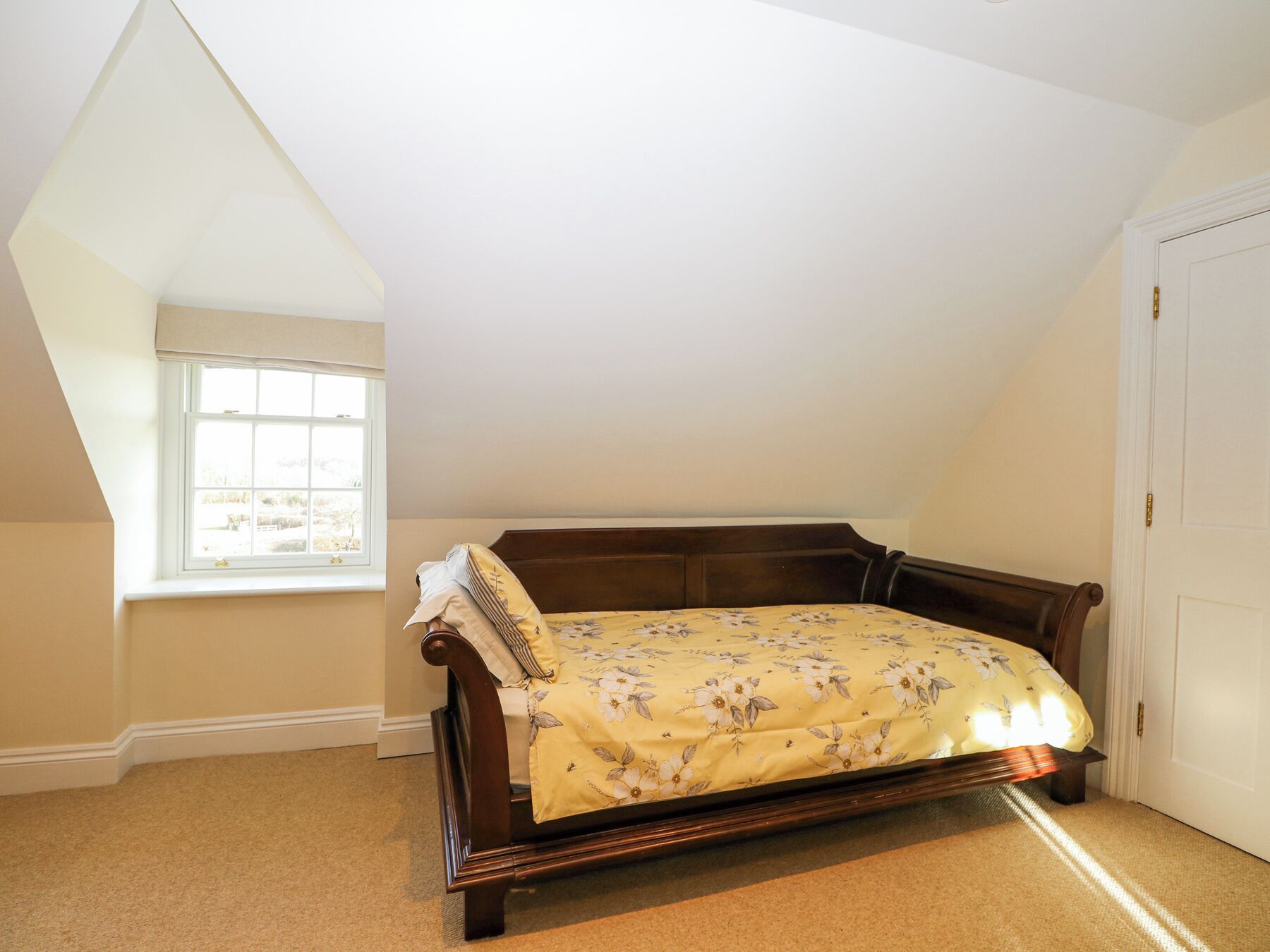Une chambre avec un plafond en pente, un lit simple recouvert d’une couvre-lit à motifs floraux, une fenêtre donnant sur un paysage et une porte.
